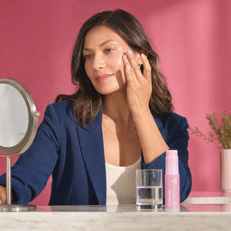 Woman in a blue blazer touching her cheek near a Yubiskin balm stick and vanity mirror on a marble surface
