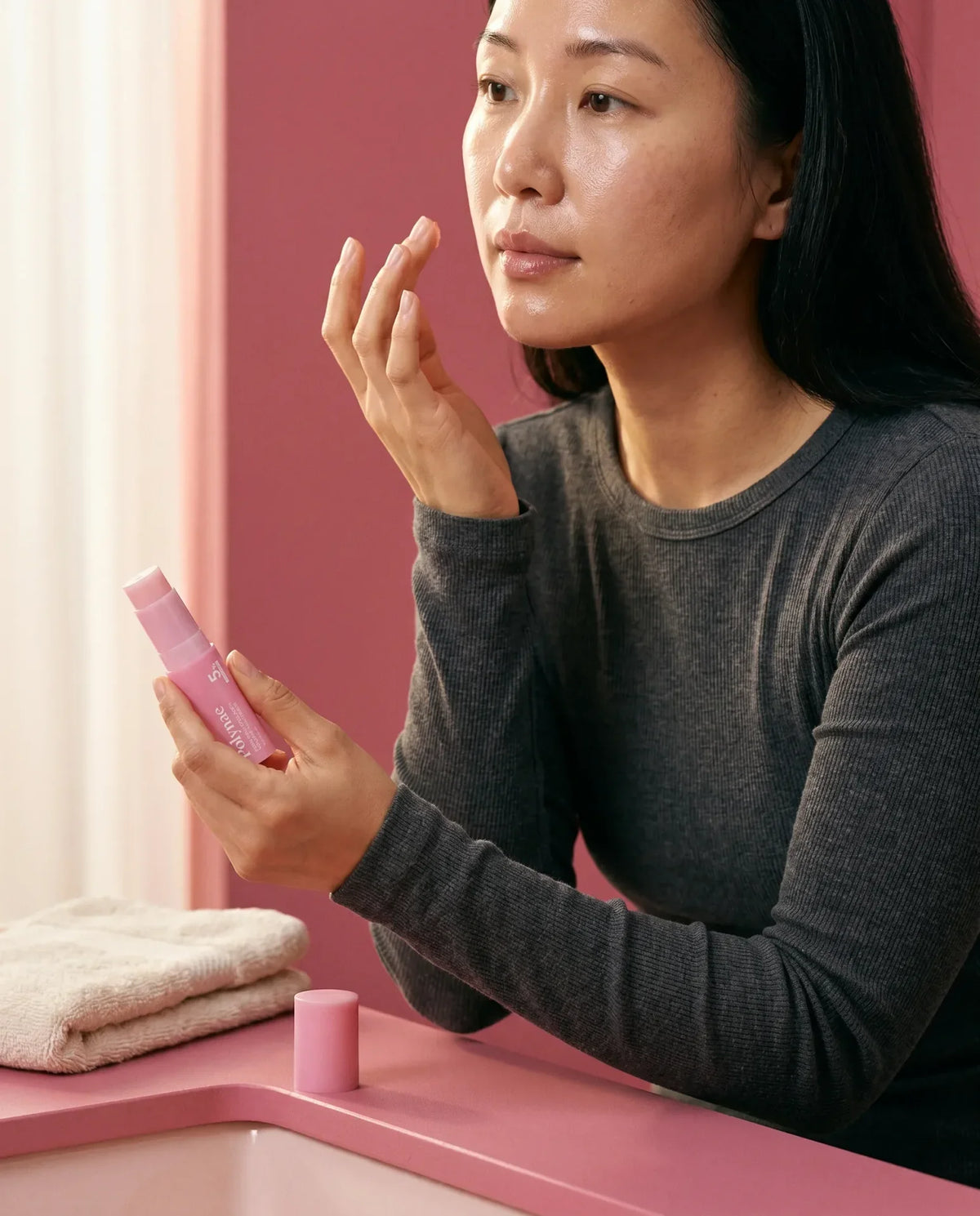 Black woman in rust turtleneck examining a pink Yubiskin product tube, reflected in a round mirror, pink background